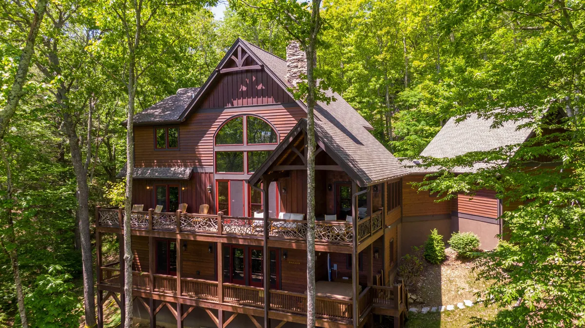This exterior shot highlights a luxury mountain home with large windows, a wooden balcony, a stone chimney, and surrounding trees. The scene conveys architectural elegance and a harmonious connection with the landscape, suggesting both retreat and community.