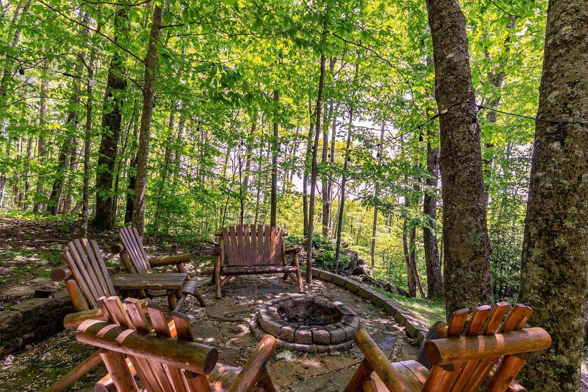 A cozy outdoor fire pit area with wooden Adirondack chairs arranged around a glowing fire, set in a natural stone seating space surrounded by trees. The scene is perfect for storytelling and stargazing in a communal, intimate setting.