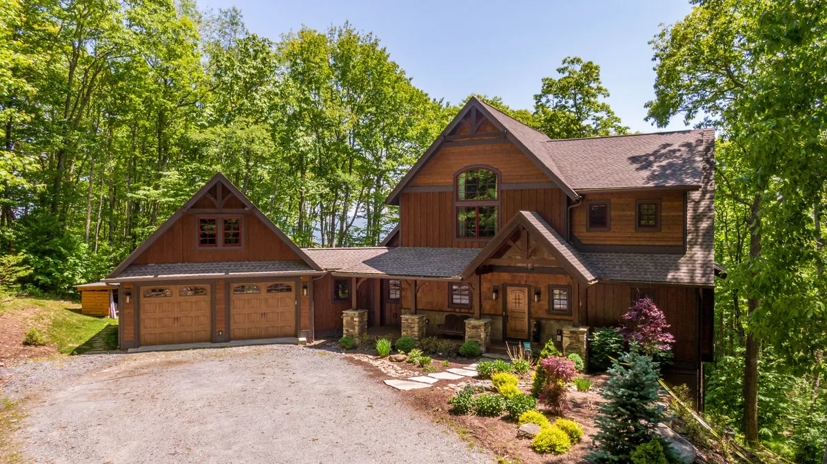 The image showcases a beautifully crafted wooden and stone single-family home nestled amidst lush trees, with a meticulously landscaped garden and spacious driveway. The inviting porch overlooks serene mountain surroundings, hinting at opportunities for sunset watching, family gatherings, and relaxing in nature.