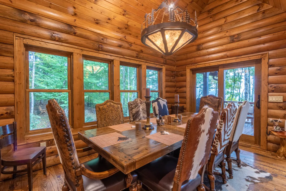 This image features a large wooden dining table beneath an ornate chandelier, surrounded by rustic wooden log walls and illuminated by natural light streaming through generous windows. The arrangement invites family feasts and celebratory gatherings while offering a view of the natural mountain backdrop.