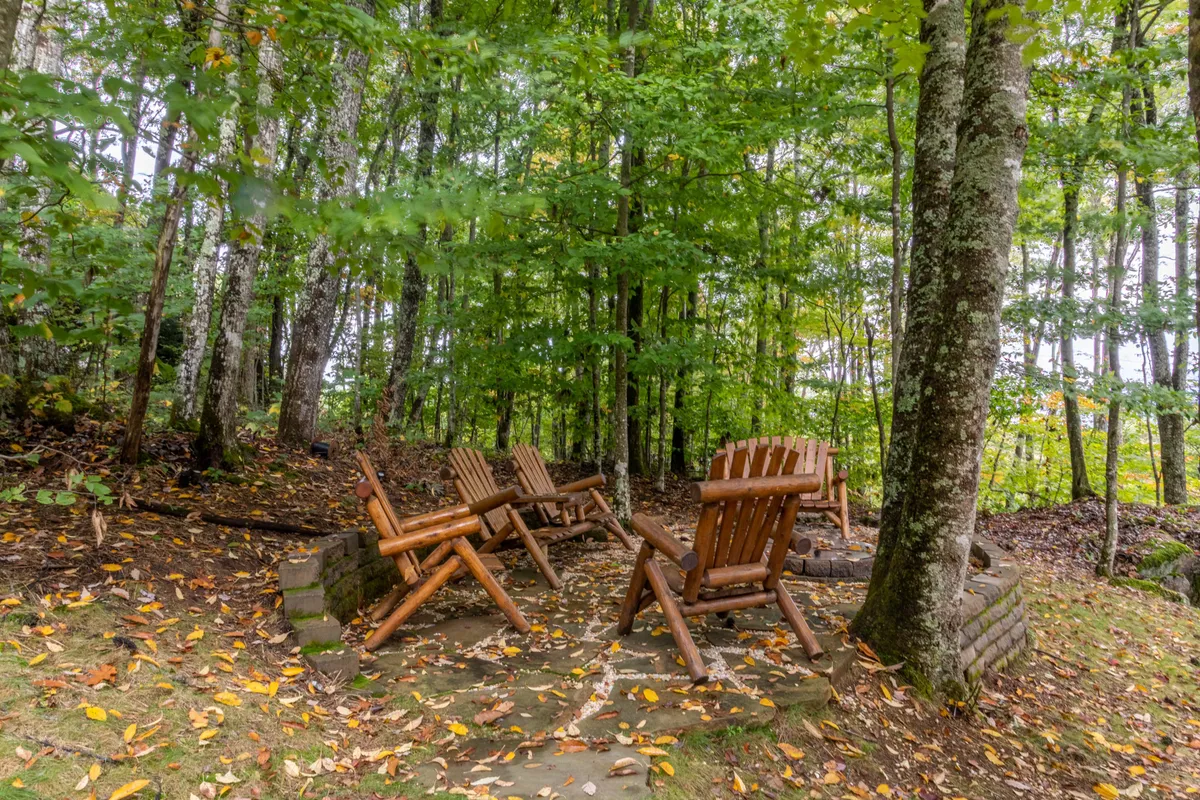 An exterior gathering spot with wooden chairs circling a sturdy stone fire pit amidst trees. The design invites families to relax, tell stories, and enjoy s’mores together under the stars, emphasizing both private enjoyment and the beauty of the natural setting.