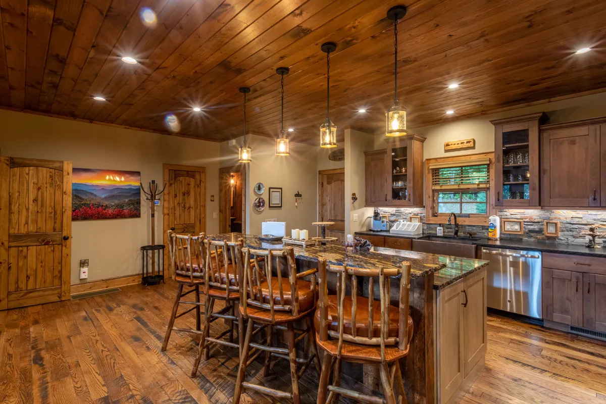 This image showcases a welcoming kitchen space featuring a kitchen island with bar stools, wooden ceiling, and a stone backsplash. The area is well-appointed for both cooking and entertaining, conveying a sense of warmth and functionality ideal for group gatherings and culinary creativity.