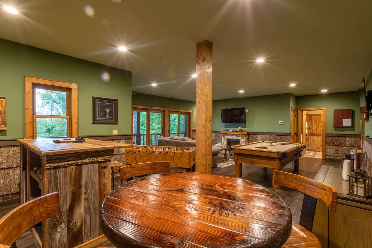 A cozy, inviting game room featuring a custom wooden pool table surrounded by comfortable seating, with large windows letting in views of lush greenery. The space is designed for social gatherings, family game nights, and quiet relaxation, and hints at a multi-use entertainment area.
