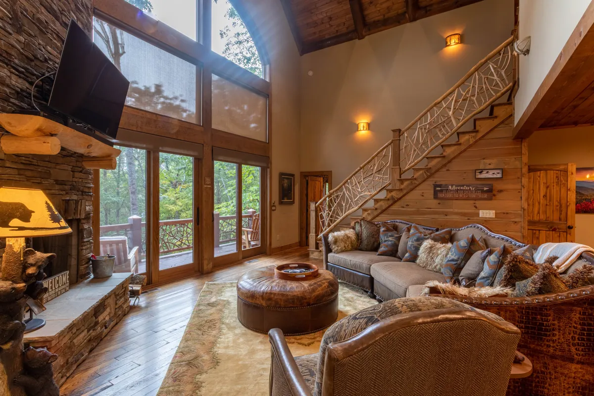 This image features a warm and welcoming living room with a dramatic stone fireplace, large windows that flood the space with natural light, and a comfortable seating area arranged for socializing or relaxing. A wooden staircase adds rustic charm and hints at the craftsmanship found throughout the home.