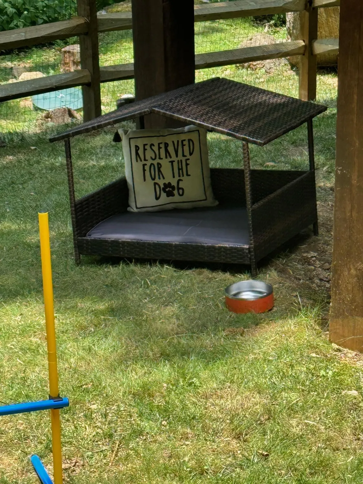 The image shows an exterior space dedicated to dogs, featuring a comfortable dog bed, a 'reserved' sign, a water bowl, and play equipment, all set under the shade. The area looks inviting for both pets and their owners, emphasizing relaxation and fun amidst natural surroundings.