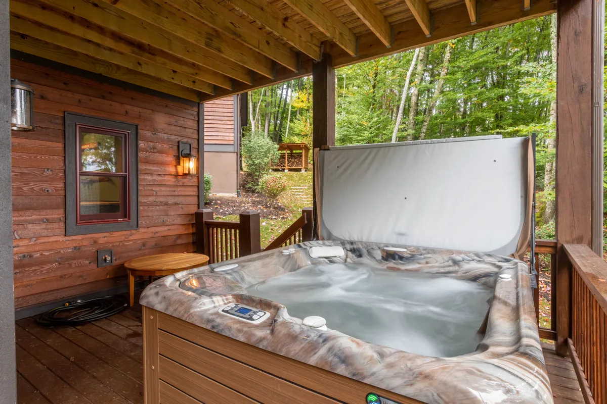 An exterior shot of a bubbling hot tub nestled on a wooden deck, surrounded by trees and ambient outdoor lighting. The setting is tranquil yet inviting, ideal for soaking under the stars or sharing laughter with friends and family.